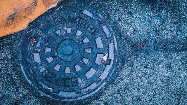 Circular metal manhole cover on a rough pavement, labeled 'Sanitary Sewer' with a radial pattern and blue tint.
