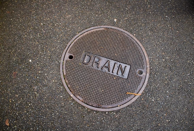 Round metal drain cover with the word 'DRAIN' embossed, set in wet asphalt surface.